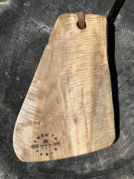 An oblong-shaped charcuterie board made of pale English Walnut with a quilted pattern on the right side. A leather strap is fixed to the top, with the Tree to Table logo engraved in the bottom.
